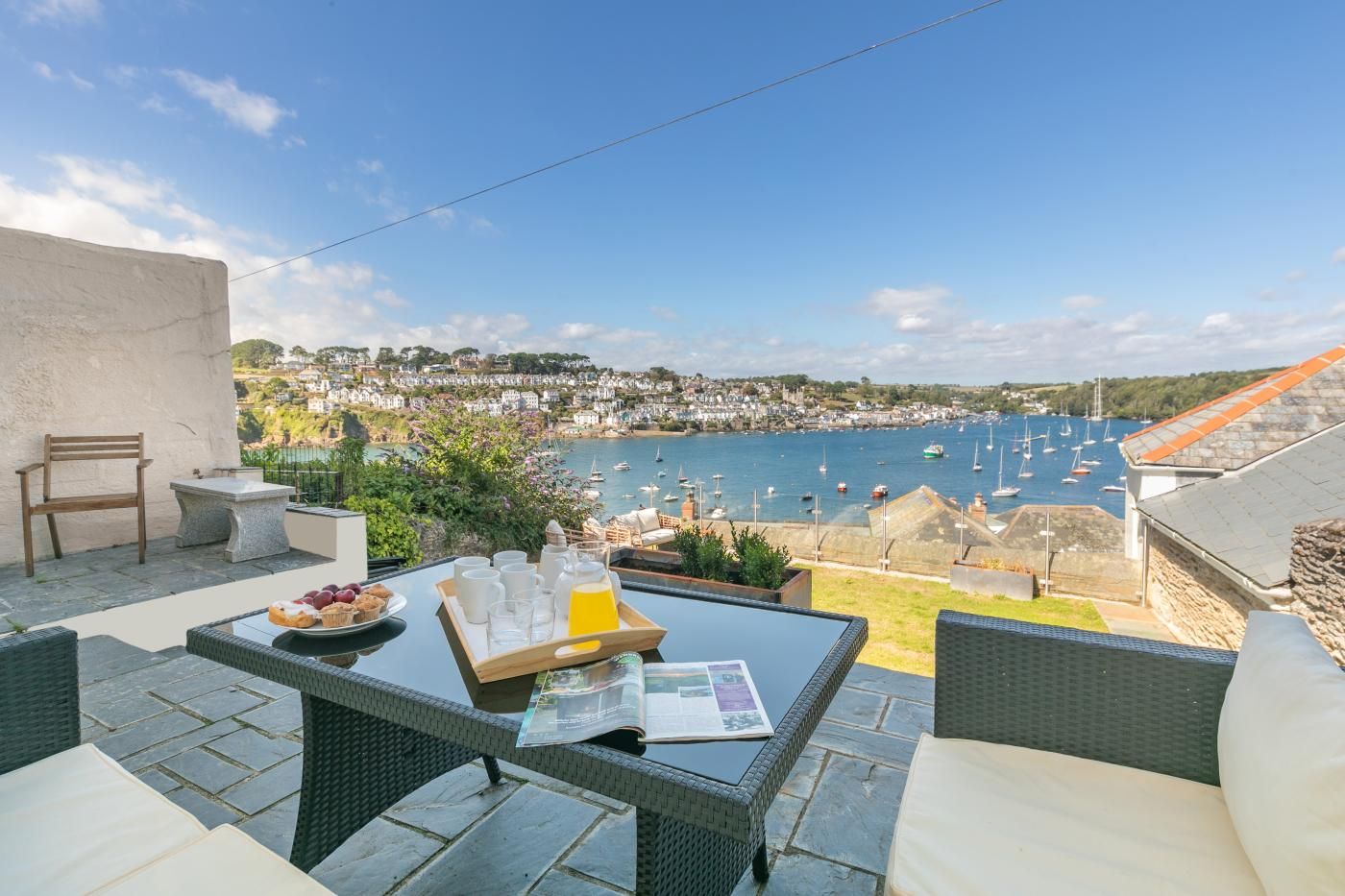 Una terraza con una mesa lista para el desayuno, con vistas a un puerto con barcos y un pueblo costero.