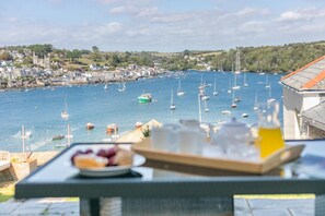 A coastal town with a marina filled with boats, a table with food and drinks, and a view of the town and hills in the background. - 3 Bed in Polruan (oc-83821) (Fowey)