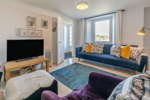 A living room with a dark blue sofa, a flat-screen TV on a wooden stand, a blue rug, and a purple armchair. - 3 Bed in Polruan (oc-83821) (Fowey)