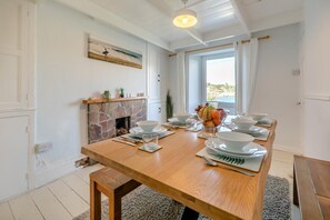 A dining room with a long wooden table set for six, a stone fireplace, and a view of the sea through a large window. - 3 Bed in Polruan (oc-83821) (Fowey)