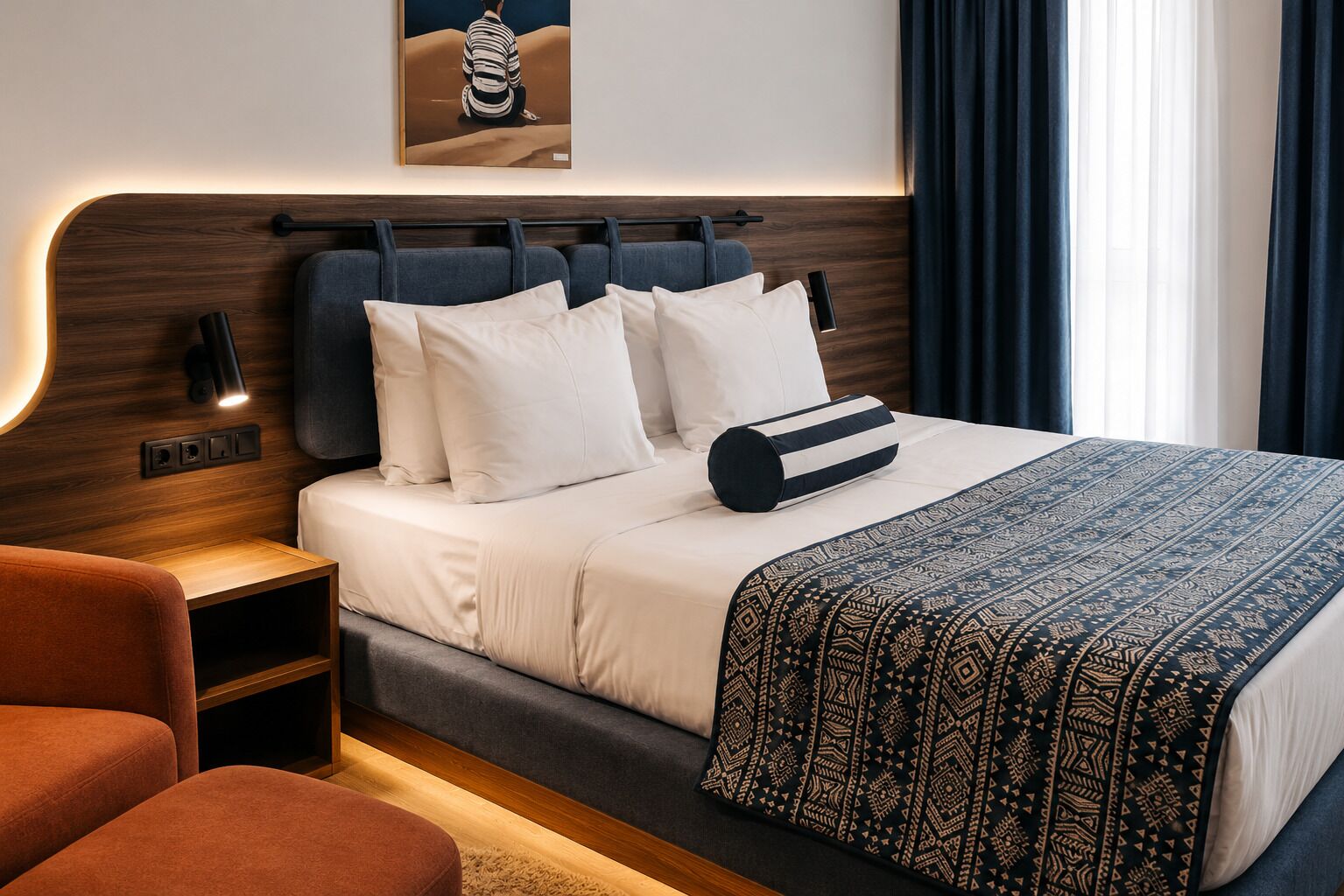 Habitación de hotel moderna con cama, mesitas de noche, lámpara de pared, manta decorativa y una obra de arte enmarcada sobre la cama.
