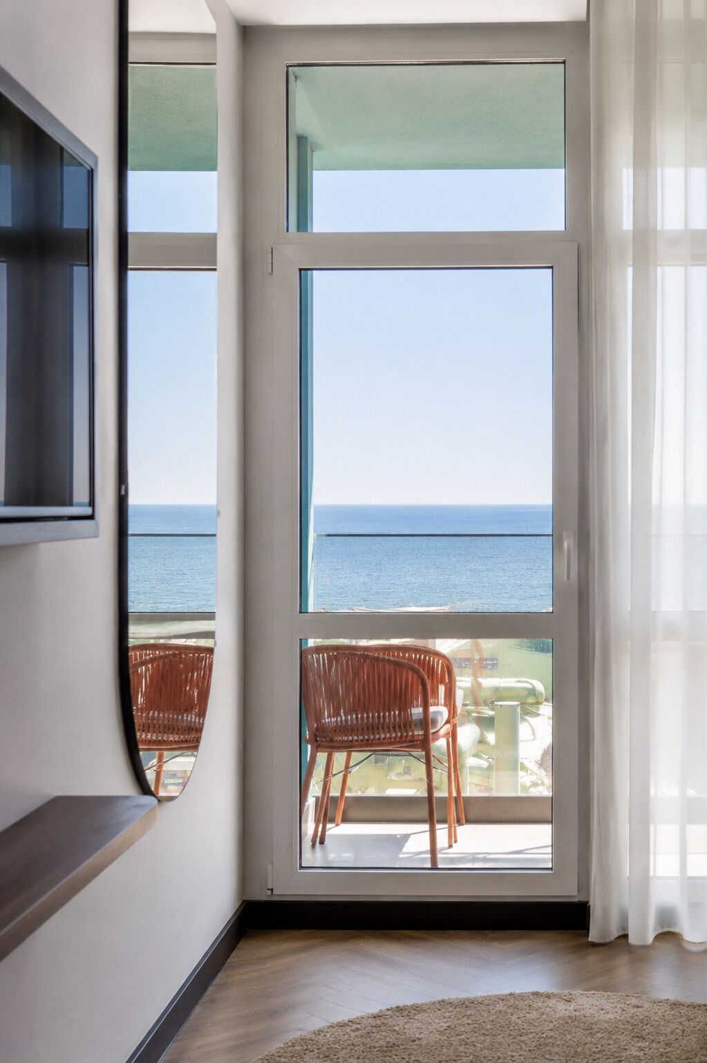 Una habitación con un ventanal grande con vista al mar, piso de madera y televisor montado en la pared.