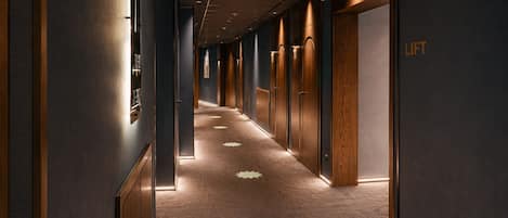 Corredor de hotel com portas de madeira, piso coberto por carpete e um aviso indicando um elevador.