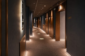 Pasillo de hotel con puertas de madera, piso alfombrado y un letrero que indica un ascensor.