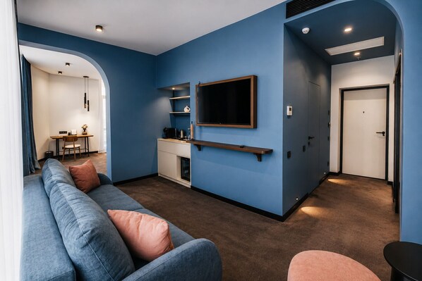 Una sala moderna con una pared azul decorativa, un televisor montado, un sofá con cojines y una zona de cocina con mostrador y armarios.