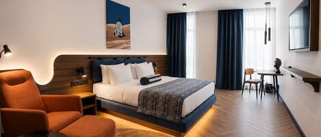 Una habitación de hotel moderna con cama, zona de estar con una silla y una mesita, escritorio con silla y un amplio ventanal con cortinas.