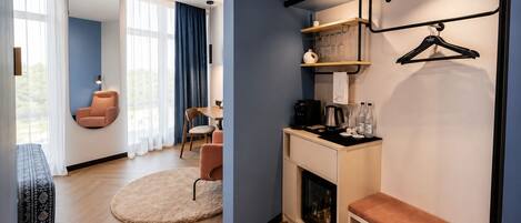A modern hotel room with a seating area, a coffee station, and a wardrobe with hanging space.