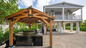 A covered outdoor area with barbecue grills, a white house with a balcony in the background. - NAUTICAL NEST - 3-bedroom canal front tranquil home on Summerland Key with AC (Summerland Key)