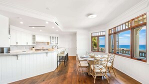 Une cuisine moderne avec des meubles blancs, un coin repas doté de chaises en osier et de grandes fenêtres offrant une vue sur la mer.