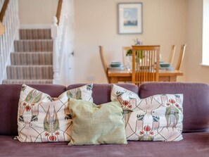 A purple sofa with patterned pillows and a plain pillow in a living room with a staircase and a dining area in the background. - 3 Bed in Bridport (oc-dc049) (Chideock)