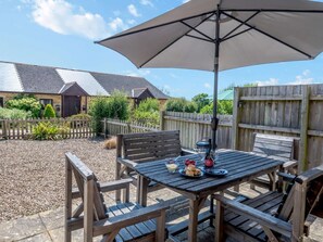 Une table de patio en bois, surmontée d’un grand parasol, entourée de chaises en bois, et une zone recouverte de gravier, avec une clôture en bois et des bâtiments en arrière-plan.