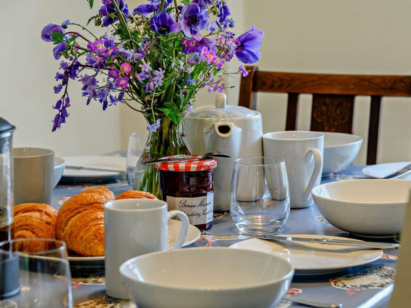 A breakfast table with croissants, a jar of jam, and a vase of flowers.