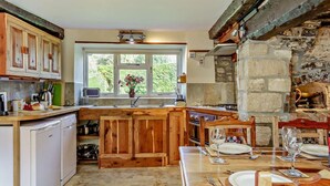 Cocina rústica con armarios de madera, pared de piedra, ventana con vistas y un comedor listo para dos personas.
