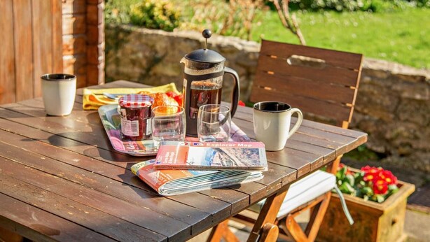 Una mesa de madera con una cafetera, una taza, un vaso, un tarro de mermelada, un libro y una revista.