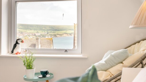 Ein gemütliches Zimmer mit Blick auf eine Küstenstadt, ein kleiner Tisch mit Pflanze und Tasse sowie ein Korbsessel mit Kissen.