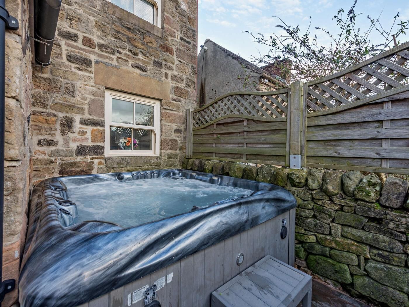 A hot tub in a stone-walled outdoor area with a wooden fence and a window.