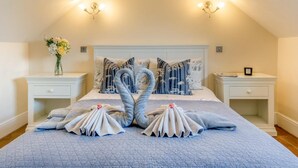 Une chambre avec un plafond en pente, un lit recouvert d’une courtepointe bleue, deux tables de chevet, un vase avec des fleurs et une décoration en forme de cœur faite avec des serviettes.