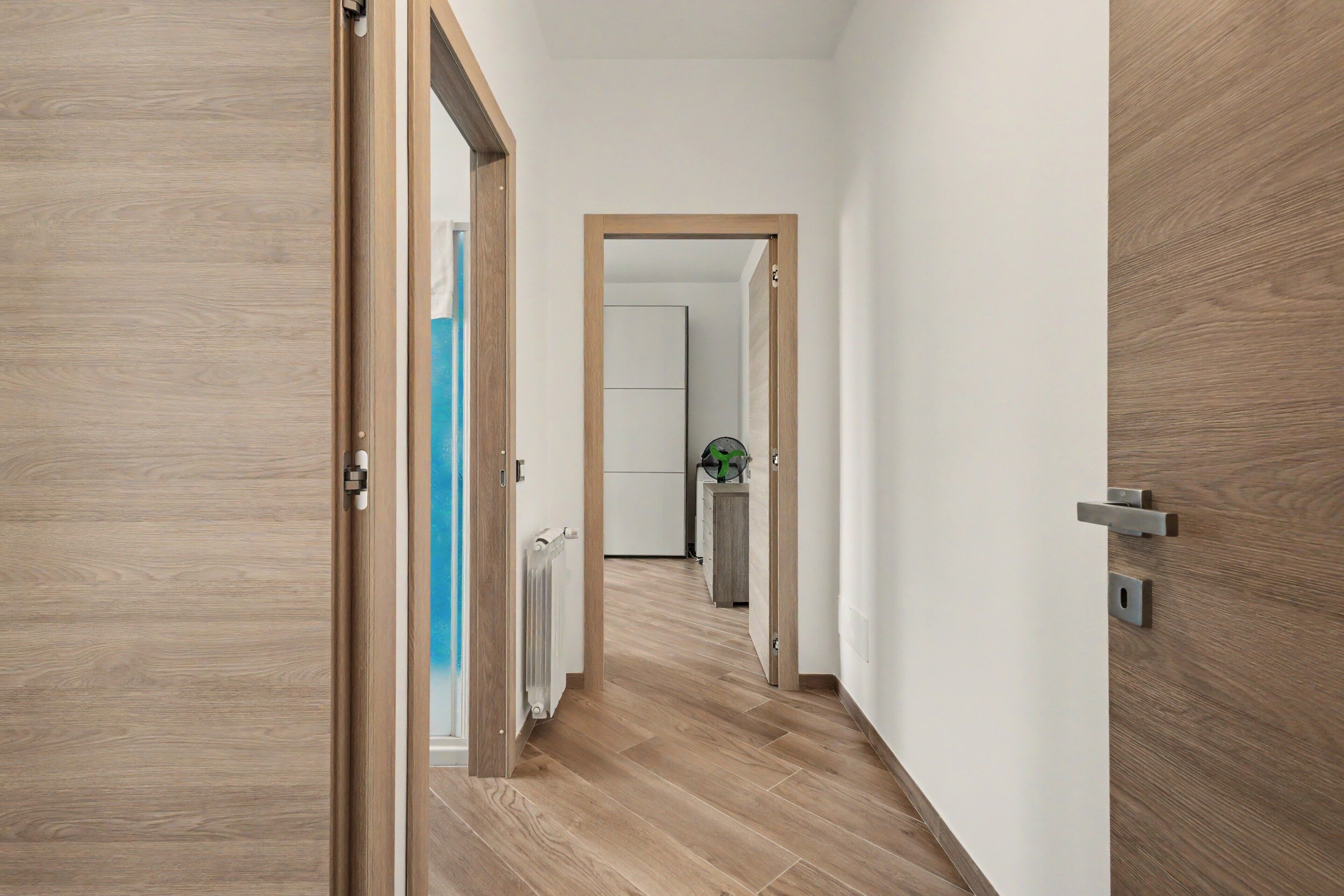 Un corridoio con pavimento in legno, un termosifone e una porta che dà su una stanza con una pianta.