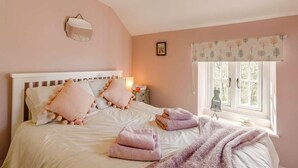Ein Schlafzimmer mit Bett, Kissen, Nachttisch, Lampe, einem Fenster mit Vorhängen und einer Wandleuchte.
