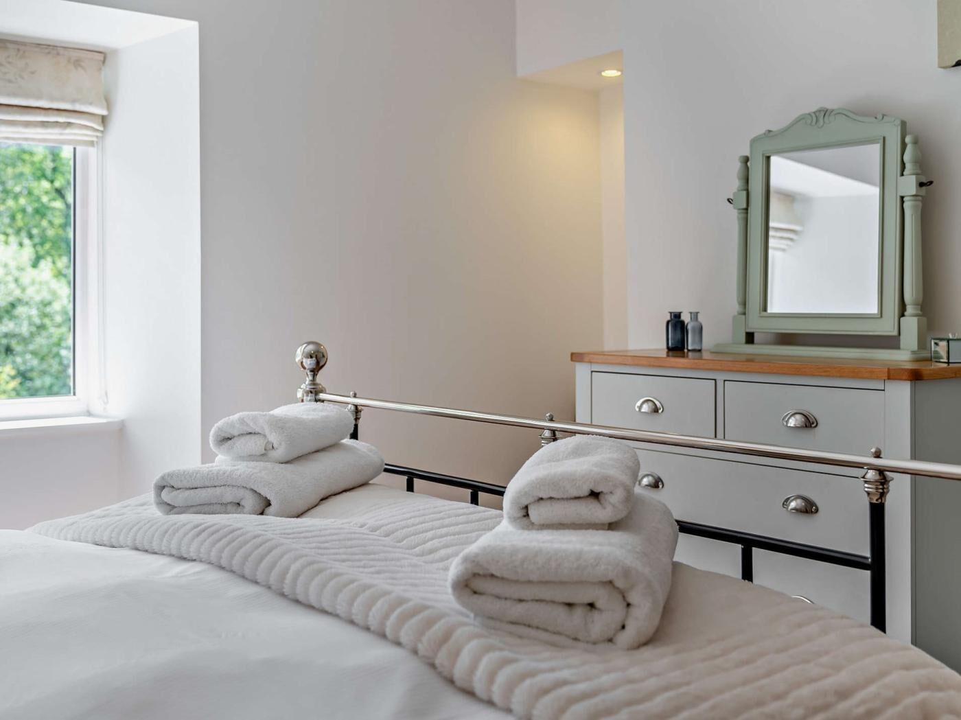 Un dormitorio con cama, mesita de noche con espejo y ventana con cortinas.