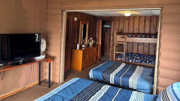 A room with wooden walls, a ceiling fan, a television on a wooden table, and two bunk beds with striped bedding.