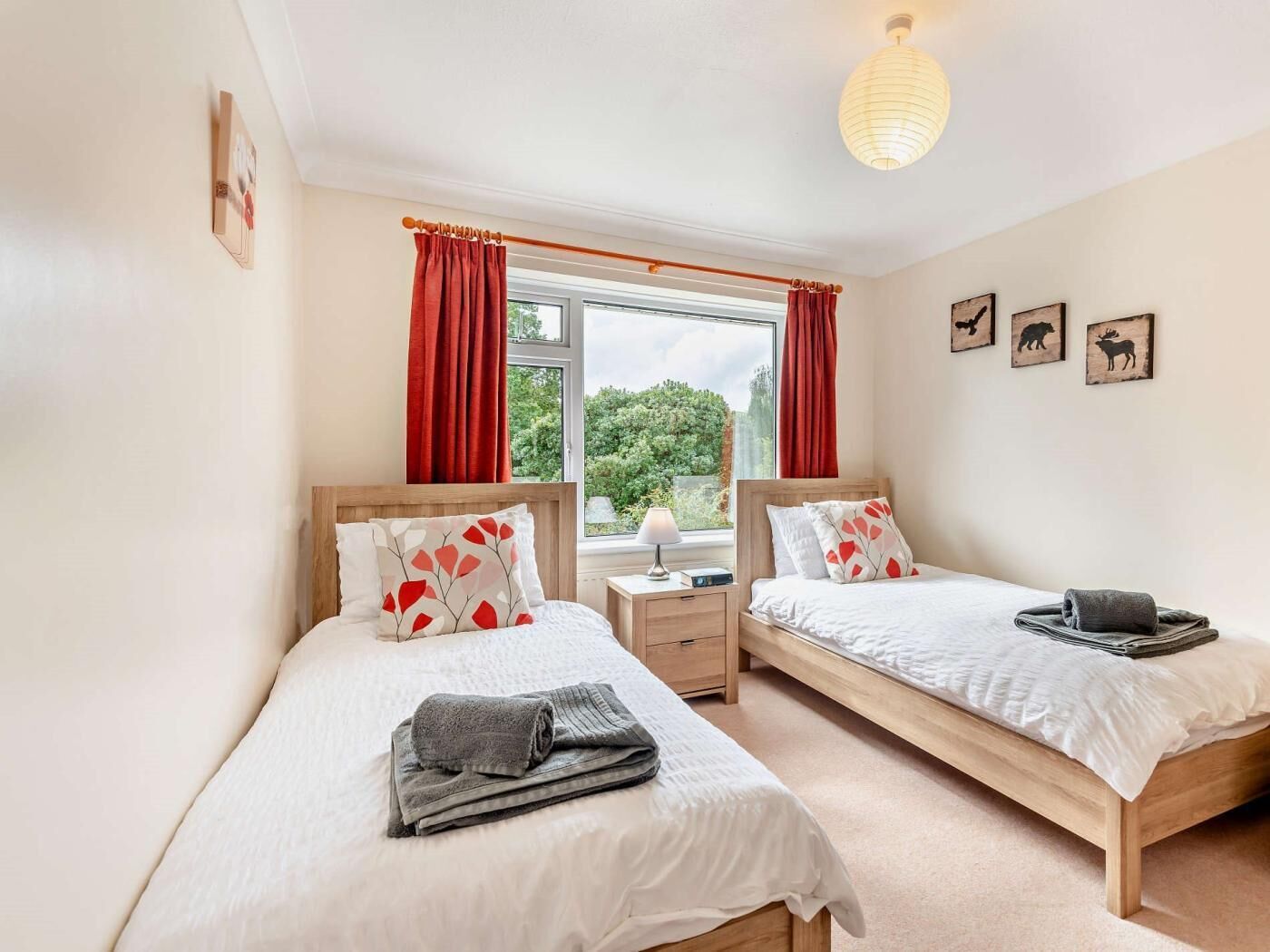 A bedroom with two beds, a window with curtains, a bedside table with a lamp, and wall art.