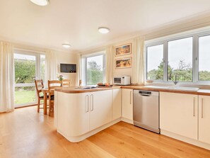 A modern kitchen with wooden cabinets, a built-in microwave, and a dishwasher. There is a dining area with chairs and a table, and large windows providing natural light. - 4 Bed in Swanage (oc-75639) (Studland)