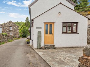 A white building with a wooden door and a window, a stone wall, and a paved driveway. - 3 Bed in Keswick (oc-sz153) (Braithwaite)