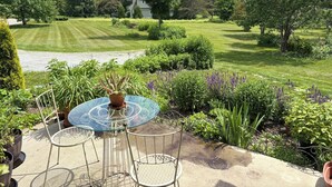 A patio with a glass table and chairs, surrounded by greenery and a view of a well-manicured lawn.
