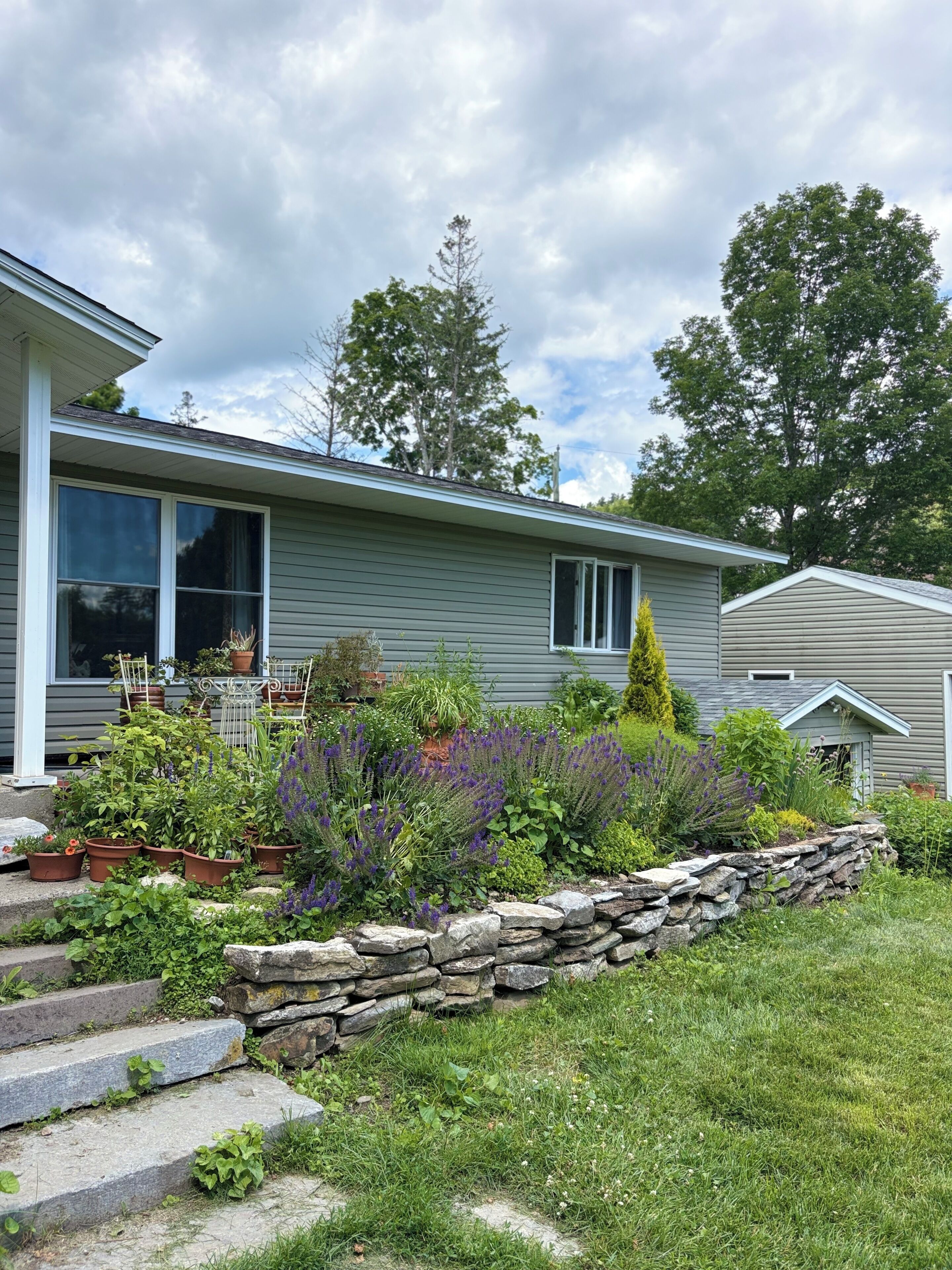 A residential house with a stone retaining wall, a front porch with potted plants, and a clear sky.