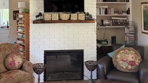 A living room with a white brick fireplace, a flat-screen TV, a ceiling fan, and a bookshelf.