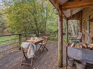 A wooden deck with a table set for two, surrounded by chairs, overlooking a forested area.