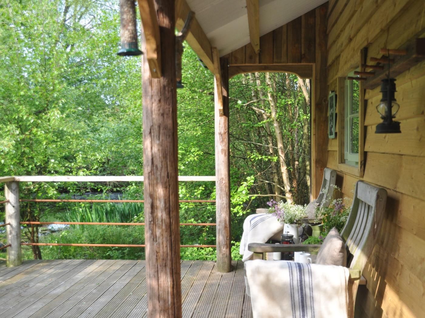 A wooden deck with a bench and table, surrounded by greenery and a wooden fence.