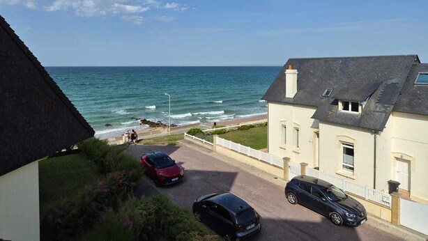 Udsigt over kysten med en vej, der fører til stranden, en rød sportsvogn og en sort bil parkeret foran en bygning.