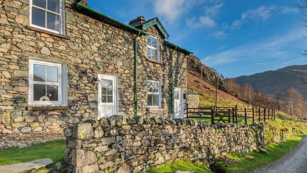Una casa de piedra con techo verde, ventanas blancas y un muro de piedra.