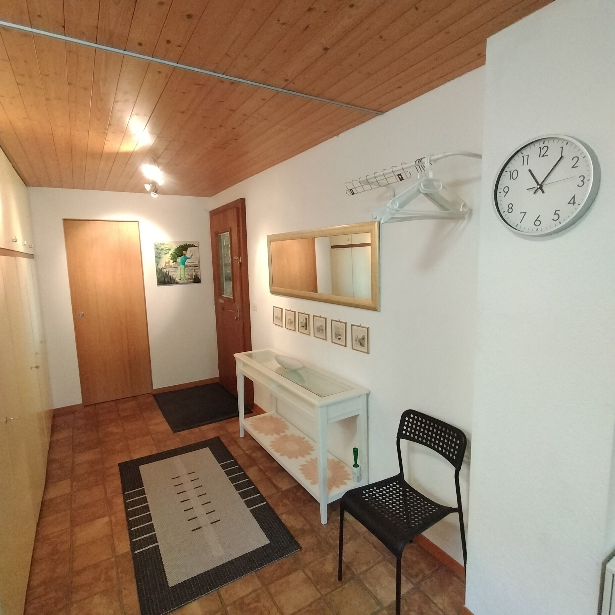 A hallway with a wooden ceiling, a white wall-mounted clock, a mirror, a white console table, a black chair, and a door.