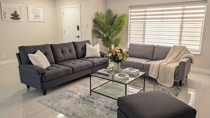 Een woonkamer met een hoekbank, een glazen salontafel, een plant in pot en een raam met zonwering.