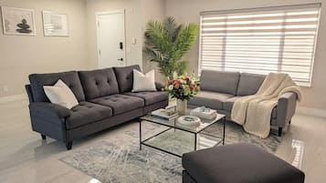 A living room with a sectional sofa, a glass coffee table, a potted plant, and a window with blinds.