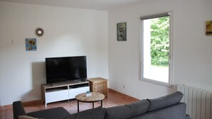 A living room with a flat-screen TV, a grey sofa, a small round table, and a large window. - Le Petit Balou Vacation Rental, Dordogne. A comfortable home near Bergerac (Lamonzie-Saint-Martin)