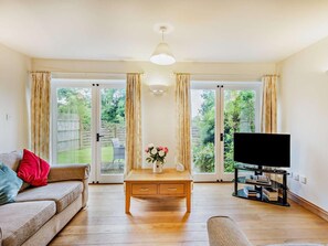 Ein Wohnzimmer mit einer Couch, einem Couchtisch mit Blumen, einem Fernseher auf einem Standfuß und einer Glasschiebetür, die in einen Garten führt.