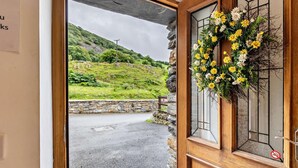 A wooden door with a stone wall, a wreath, and a view of a green landscape. - 3 Bed in Arthog (oc-77304) (Arthog)