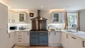 A modern kitchen with white cabinetry, a stainless steel range hood, a microwave, a toaster, and a window with curtains. - 3 Bed in Arthog (oc-77304) (Arthog)