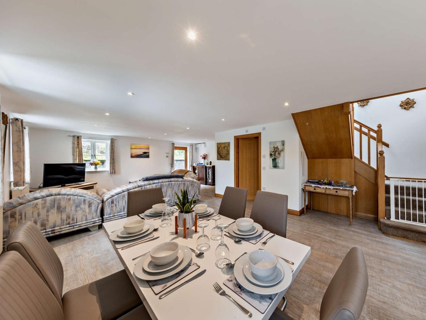 A modern dining area with a large table set for four, a sofa, and a staircase leading to an upper level.