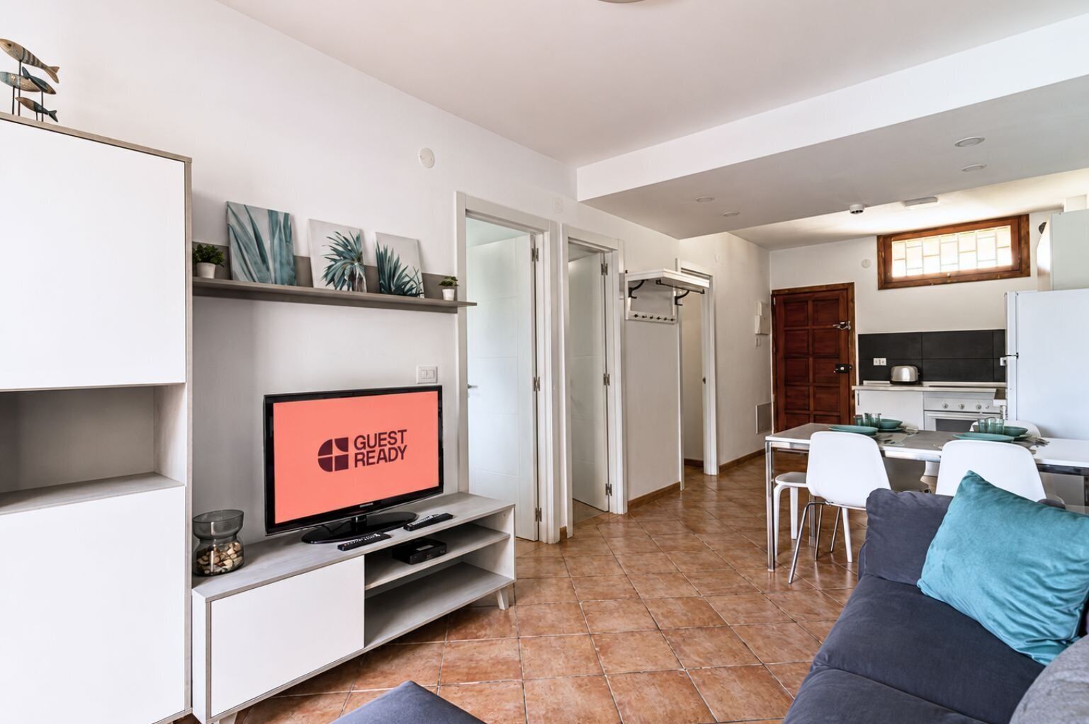 A modern living room with a television displaying 'GUEST READY,' a dining area with a table and chairs, and a kitchen with a stove and refrigerator.