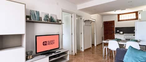 Una sala de estar moderna con un televisor que muestra 'GUEST READY', un comedor con mesa y sillas, y una cocina con estufa y refrigerador.