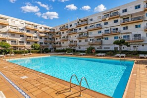 Ein Schwimmbecken mit gefliestem Rand, umgeben von einem mehrstöckigen Gebäude mit Balkonen und blauem Himmel.