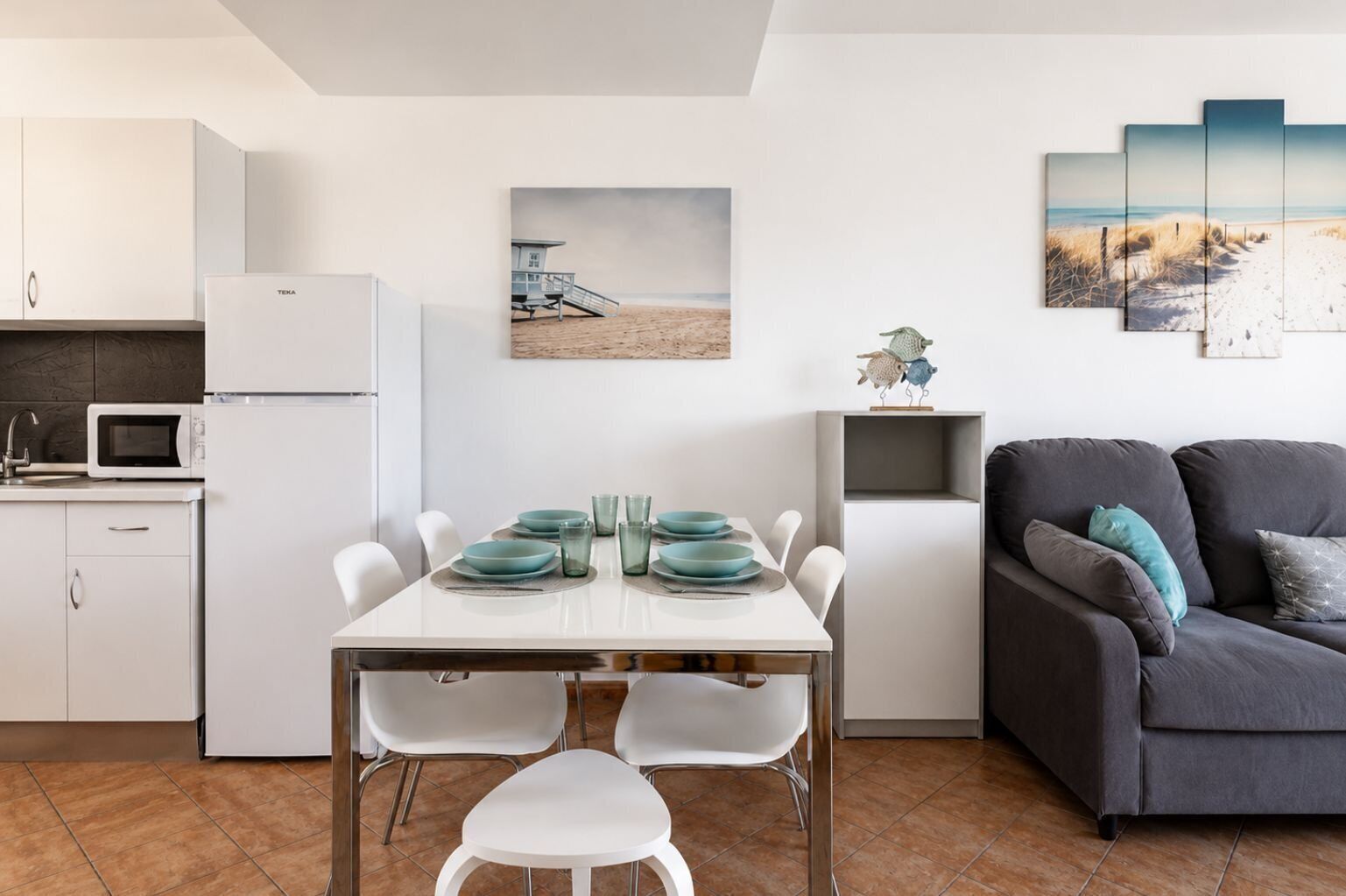 A modern kitchen with white cabinets and a microwave, a dining area with a white table and teal plates, and a gray sofa with teal cushions.