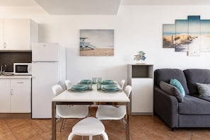 A modern kitchen with white cabinets and a microwave, a dining area with a white table and teal plates, and a gray sofa with teal cushions. - GuestReady - Tenesor Sun Beach Resort (Las Palmas de Gran Canaria)