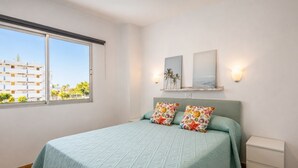 A bedroom with a bed, two pillows, a nightstand, a window, and wall-mounted artwork. - GuestReady - Tenesor Sun Beach Resort (Las Palmas de Gran Canaria)