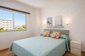 A bedroom with a bed, two pillows, a nightstand, a window, and wall-mounted artwork. - GuestReady - Tenesor Sun Beach Resort (Las Palmas de Gran Canaria)